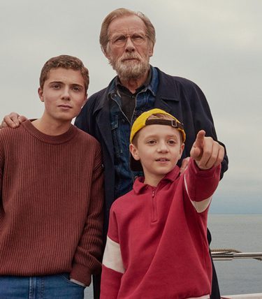 Bill Nighy is with two young boys on a boat, one of the boys is pointing off to the sea.