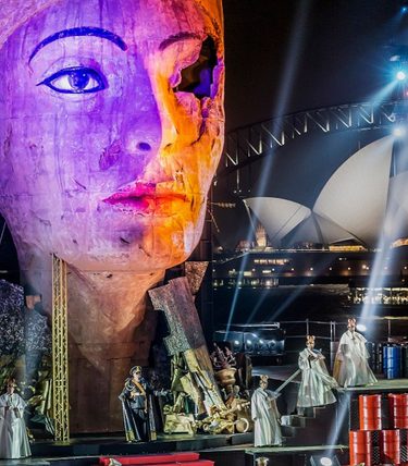 A wide shot of an opera stage featruing many performers a huge head prop and the Sydney Opera House seen in the background.