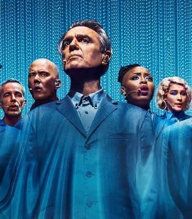 David Byrne stands centrally with his cast all dressed in blue against a blue background.