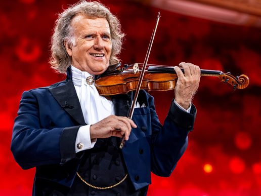 Andre Rieu is stood with his violin under his chin. He is against a red backdrop.