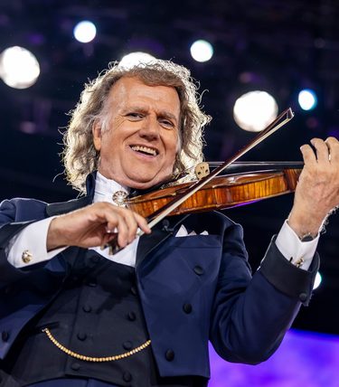 Andre Rieu smiles at the camera while playing his violin live on stage.