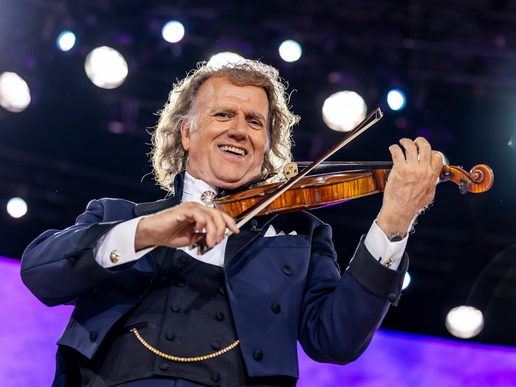 Andre Rieu smiles at the camera while playing his violin live on stage.