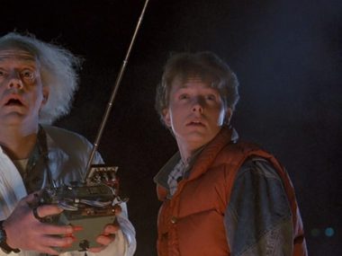 A young man and an older scientist are stood next to each other on a dark night. The scientist has a large remote control.