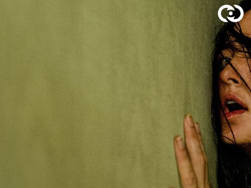 A woman is pressed up against a yellow wall. She is gasping in fear.