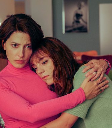 Two women are sat on a sofa with one hugging the other.