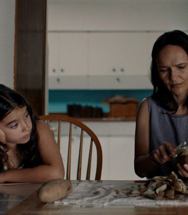 A young girl is sat at a table with a woman who looks to be teaching her something.
