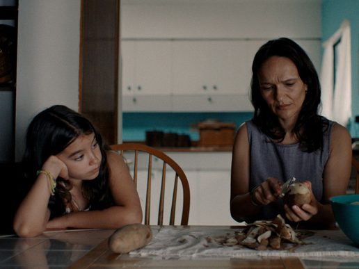 A young girl is sat at a table with a woman who looks to be teaching her something.