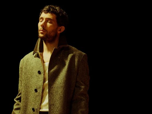 Josh O'Connor stands against a black background in a brown jacket with his eyes closed contemplatively.
