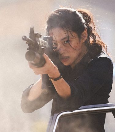 A woman is leaning out of a car with a huge gun aimed at something off screen.