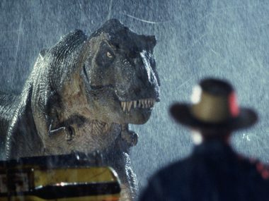 A dark rainy night with a T-rex looking toward a man holding a flare.