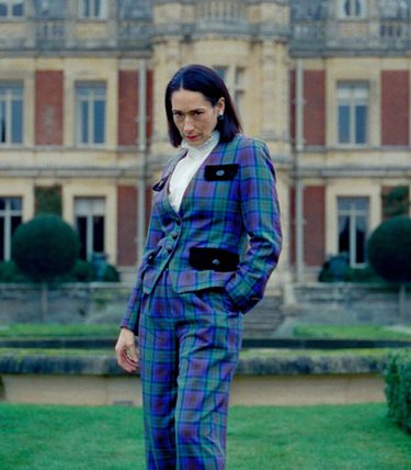 An aristocratic lady in a striking blue suit is outside a stately manor.