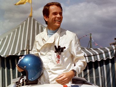 A racing driver stands over a car clutchign his helmet. He is in front of some tents.