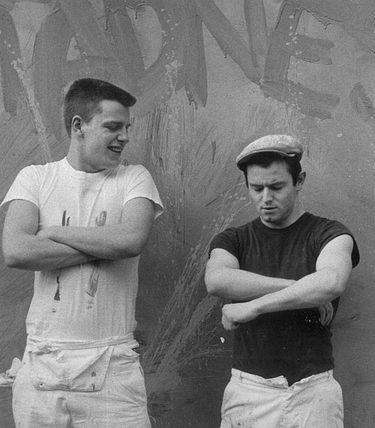 Two members of Madness in a black and white photo against aa wall where they have painted 'Madness'.