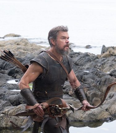 Matt Damon is in medieval garb with a bow and arrow, running across a stony beach.
