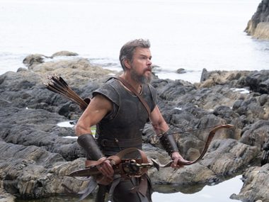 Matt Damon is in medieval garb with a bow and arrow, running across a stony beach.