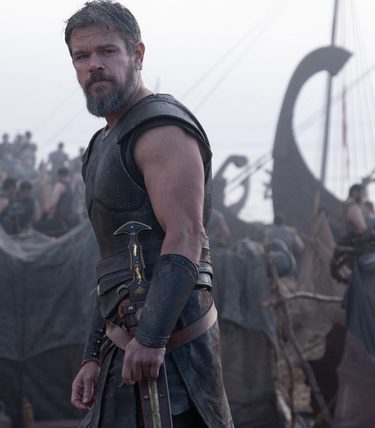 Matt Damon stands with a sword in front of a longship and surrounded by other crew members.