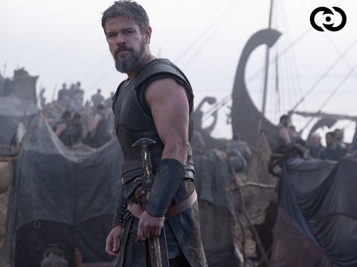 Matt Damon stands with a sword in front of a longship and surrounded by other crew members.
