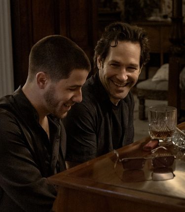 Paul Rudd and Nick Jonas are both sat at a piano and smiling. There are glasses of whickey on top.