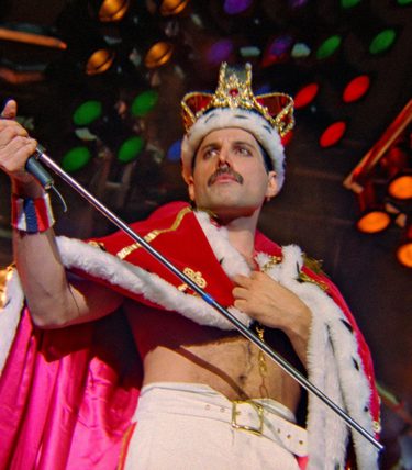 Queen singer Freddie Mercury is wearing a crown onstage in a defiant pose.