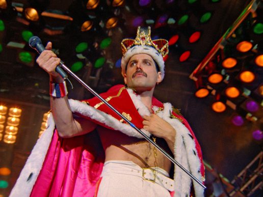 Queen singer Freddie Mercury is wearing a crown onstage in a defiant pose.