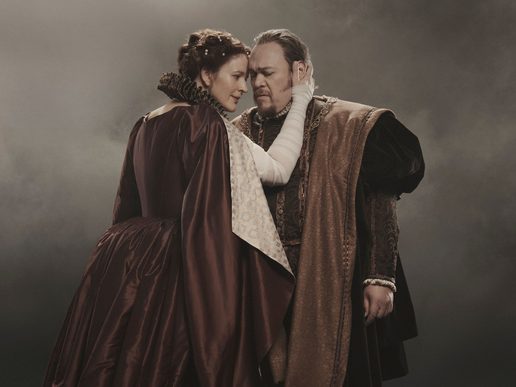 A man is stood solemnly while a woman looks into his eyes and embraces him. A misty background is behind them.