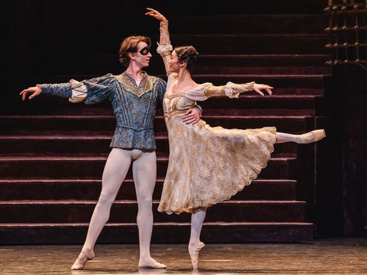 A young man and woman are dancing together. The man is masked, the woman is in a flowing, flowery dress.