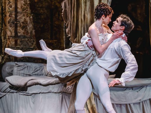 A female dancer is being held horizontally by a male dancer, both dressed in white.
