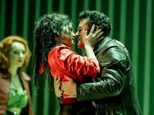 A man and woman are locked in a passionate embrace against a green curtained backdrop with other singers in the background.