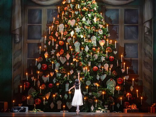 A huge Christmas tree covered in lights and baubles occupies most of the image with a young ballet dancers standing in front of it.