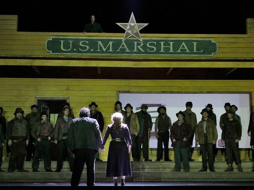 A large ensemble of performers are all pointing guns at a cast member on the right.