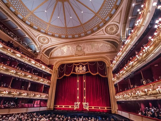 An internal view of the Royal Opera House with a full audience. Opulent gold trim around the fixtures and a velvet red curtain.