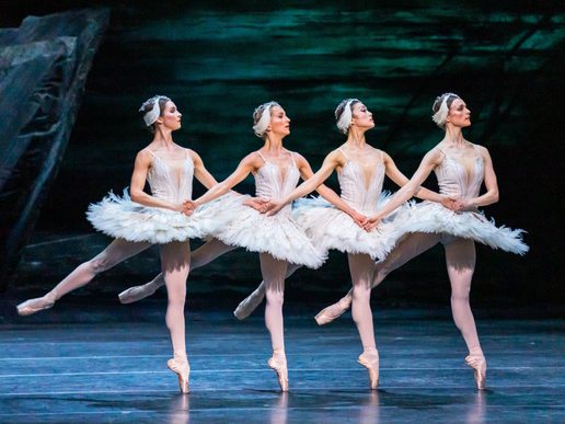 Four female ballet dancers in traditional white clothing are joining hands and on tip-toes, mid-movement.