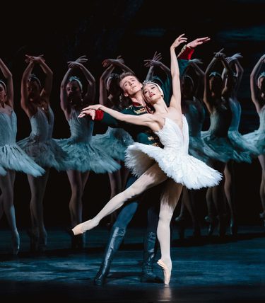 A male and female ballet dancers are mid-dance in front of a chorus line of dancers.