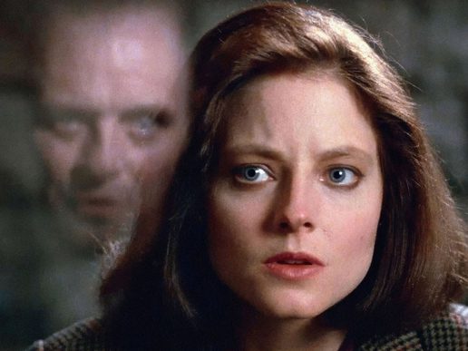 A womans face is in the foreground, she is talking to a man who is seen reflected in the glass between them.