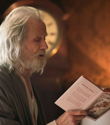Bill Nighy is in profile reading from a book. He has long white hair and appears to be in a dressing gown.
