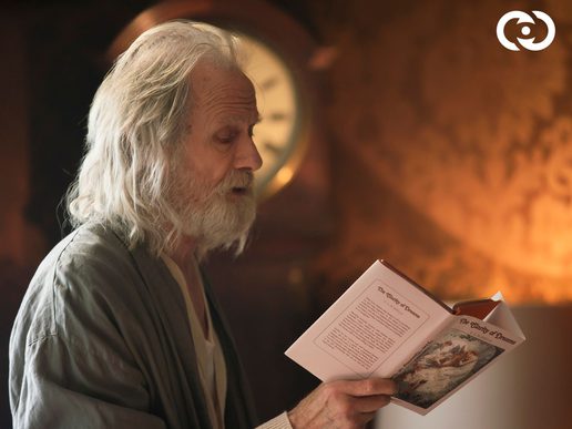 Bill Nighy is in profile reading from a book. He has long white hair and appears to be in a dressing gown.