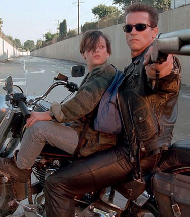 Arold Schwarzenegger on a motorbike with a teenage boy. He is holding a shotgun toward an offscreen villain.