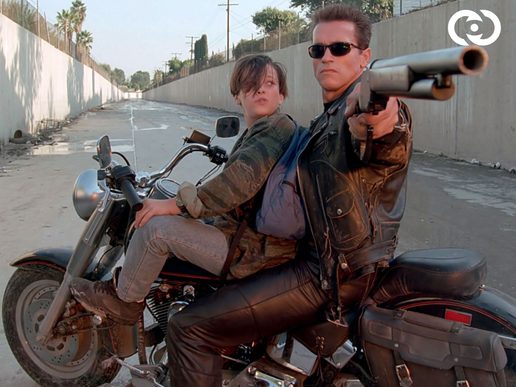 Arold Schwarzenegger on a motorbike with a teenage boy. He is holding a shotgun toward an offscreen villain.
