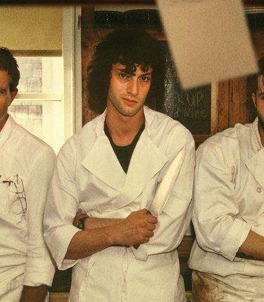 Three men in Chefs whites are standing in a line posing for a photo. An older one looks smart while two younger men look disheveled.