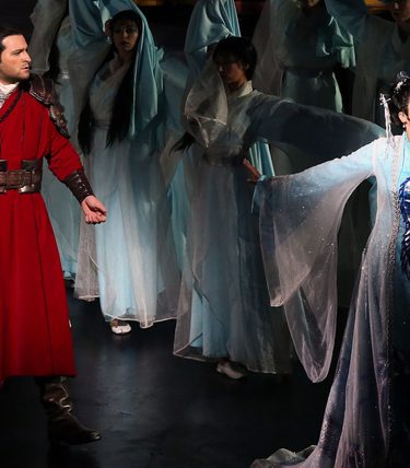 Two performers are surrounded by an ensemble. A man in a red cloak and a woman in a white outfit.