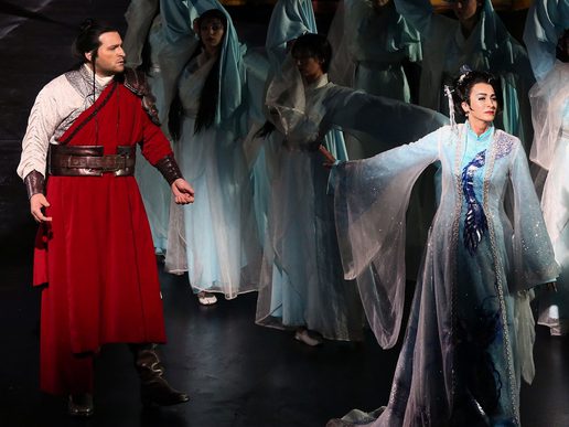 Two performers are surrounded by an ensemble. A man in a red cloak and a woman in a white outfit.
