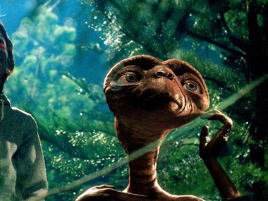 A young boy is standing next to a short brown alien in the forest, they are both looking toward the sky.