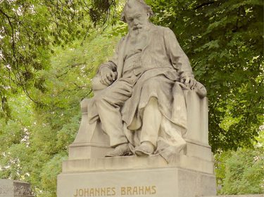A stone statue of Johannes Brahms, depicted seated with a long beard and flowing robes, mounted on a pedestal inscribed with his name, surrounded by green trees.