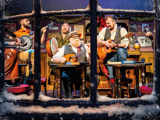 Four male presenting musicians perform traditional Irish music inside a cosy pub, seen through a frosted window with snow along the sill. They sit on stools playing banjo, drum, and guitars, smiling and mid-song, with festive garlands, vintage posters, and warm lighting creating a lively, intimate atmosphere.