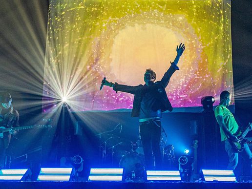 The band performs on stage, with the lead singer raising their arm in the air while holding a microphone, silhouetted against a bright starburst light and a colorful swirling backdrop, flanked by guitarists on either side.
