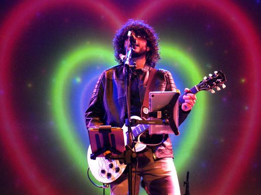 A musician with curly dark hair sings into a microphone while holding a guitar, performing on stage in front of a glowing heart-shaped light display in shades of pink, green, and purple.