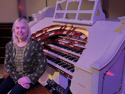 Elizabeth Harrison seated beside a white Wurlitzer theatre organ, featuring multiple keyboards and rows of illuminated controls.