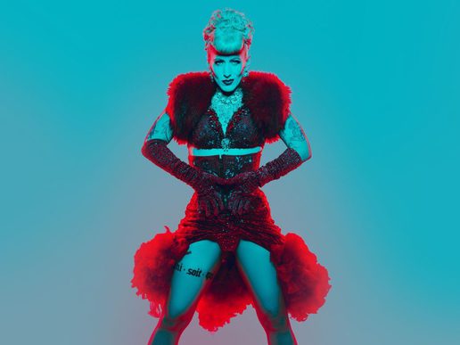 A burlesque performer poses confidently against a vivid turquoise background, illuminated by dramatic red and blue lighting. Wearing a sparkling black corset dress with a deep red feathered collar, glittering elbow-length gloves, and statement jewellery, they stand with hands on their waist and gaze directly at the camera. Their platinum-blonde hair is styled in an elaborate vintage updo, while visible tattoos and theatrical makeup add to the bold, glamorous cabaret aesthetic.