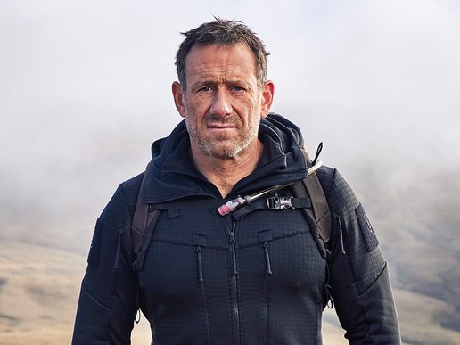 Jason Fox wearing a dark outdoor jacket and backpack, standing outdoors with a misty, mountainous landscape in the background.