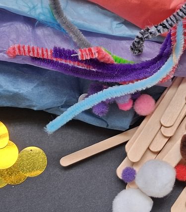 A colourful assortment of children's craft materials including pastel tissue paper, pipe cleaners, lolly sticks, pom-poms, and shiny yellow sequins, laid out on a dark surface—used in the Seaside Crafts kids workshop.